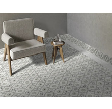 Ljust rum med mönstrat klinkergolv och matchande väggbård, en grå fåtölj och ett litet sidobord i trä.