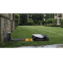 Robotgräsklippare Mammotion på en grön gräsmatta som kör mot laddstationen i regn framför en stenvägg.