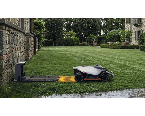 Robotgräsklippare Mammotion på en grön gräsmatta som kör mot laddstationen i regn framför en stenvägg.