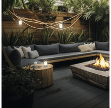 Modern terrass med L-soffa, kuddar, eldstad, träbord och bärbara LED-lampor bland växter och en trävägg.