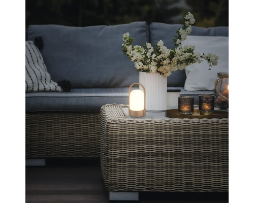 Bärbar LED-lykta på ett soffbord i konstrotting i en loungedel för trädgården med soffa, kuddar och en vas med blommor.
