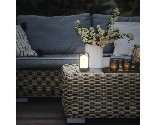Bärbar LED-bordslampa för utomhusbruk på ett rottingbord i en varmt belyst trädgårdslounge med soffa, kuddar och en vas med blommor.