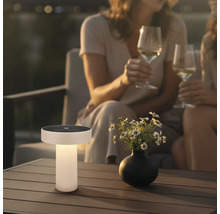 Modern solcellsbordslampa på ett terrassbord i trä i en trädgård vid skymning med blommor och personer i bakgrunden.