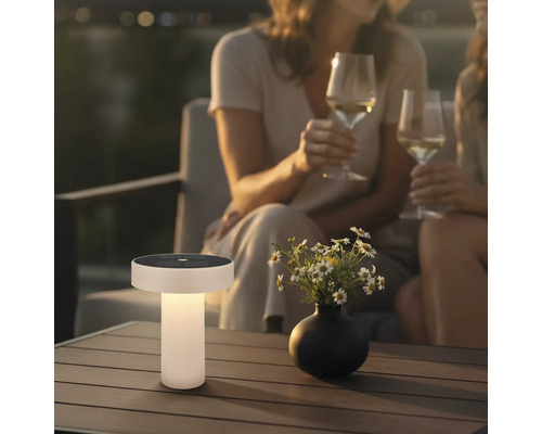 Modern solcellsbordslampa på ett terrassbord i trä i en trädgård vid skymning med blommor och personer i bakgrunden.