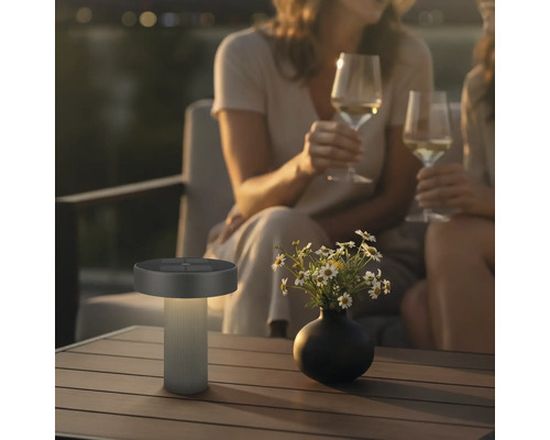 Modern LED-solcellsbordslampa för utomhusbruk på ett träbord i en mysig kvällsträdgård med människor.