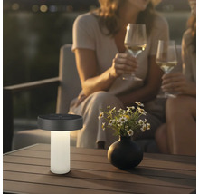 Modern solcellsbordslampa på ett terrassbord bredvid en blomvas, i bakgrunden två personer med vinglas i kvällsatmosfär.