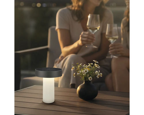 Modern solcellsbordslampa på ett terrassbord bredvid en blomvas, i bakgrunden två personer med vinglas i kvällsatmosfär.