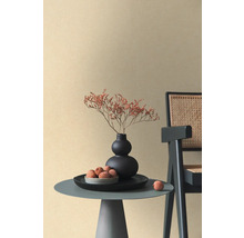 Beige non-woven tapet med fin textur i ett minimalistiskt rum med sidobord, svart vas och rottingstol.