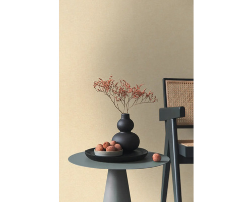 Beige non-woven tapet med fin textur i ett minimalistiskt rum med sidobord, svart vas och rottingstol.