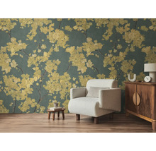 Non-woven fototapet med gula blomgrenar på mörk bakgrund i ett vardagsrum med ljus fåtölj och trämöbler på laminatgolv.