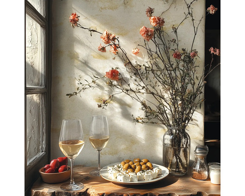 Rustikt bord vid fönstret med vitt vin, oliver, fetaost, tomater och en vas med torkade blommor framför en strukturerad vägg.