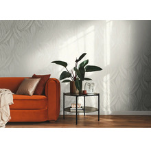 Ljus nonwoven-tapet med grafiskt bladmöster i ett modernt inrett vardagsrum med orange soffa, sidobord och inomhusväxt.
