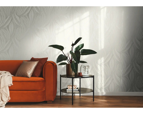 Ljus nonwoven-tapet med grafiskt bladmöster i ett modernt inrett vardagsrum med orange soffa, sidobord och inomhusväxt.