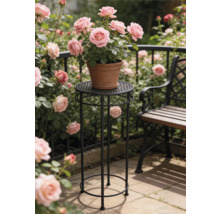 Högt runt blompiedestal i svart metall med ornament med en lerkruka med rosa rosor i en ljus trädgård bredvid en bänk.