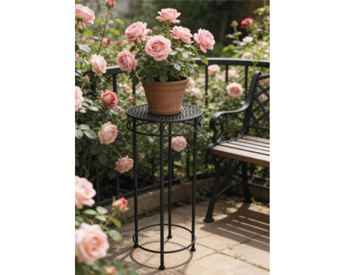 Högt runt blompiedestal i svart metall med ornament med en lerkruka med rosa rosor i en ljus trädgård bredvid en bänk.