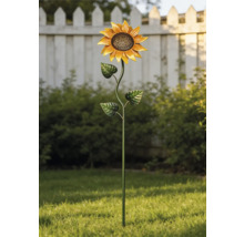 Trädgårdsdekoration i metall formad som en solros i en ljus trädgård på en grön gräsmatta framför buskar och ett vitt staket.