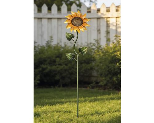 Trädgårdsdekoration i metall formad som en solros i en ljus trädgård på en grön gräsmatta framför buskar och ett vitt staket.