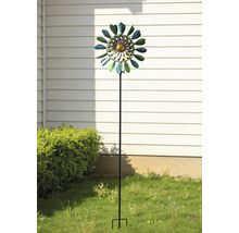 Vindsnurra i metall i form av en blomma på en trädgårdsstav, placerad i en trädgård framför en husvägg och en buske.