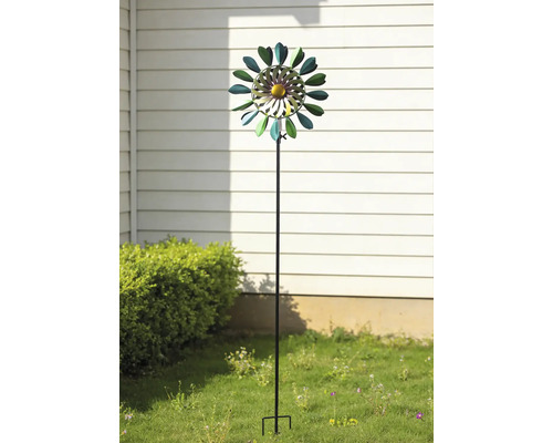 Vindsnurra i metall i form av en blomma på en trädgårdsstav, placerad i en trädgård framför en husvägg och en buske.
