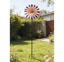 Färgglad vindsnurra av metall på en svart stång, placerad i en ljus trädgård på en gräsmatta.