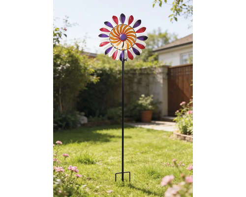 Färgglad vindsnurra av metall på en svart stång, placerad i en ljus trädgård på en gräsmatta.