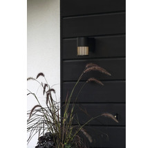 Modern svart utomhusvägglampa med vertikala ljusslitsar på en mörk trävägg bredvid prydnadsgräs.