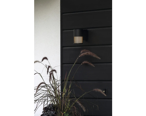 Modern svart utomhusvägglampa med vertikala ljusslitsar på en mörk trävägg bredvid prydnadsgräs.