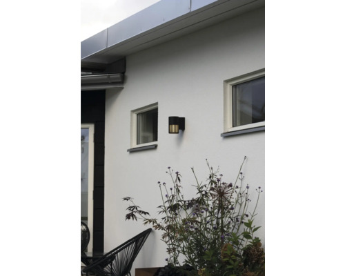 Modern svart cylindrisk utomhusvägglampa med vertikala ljuslameller på en vit husvägg mellan fönster och växter.