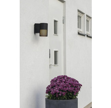 Modern svart utomhusvägglampa på en vit fasad bredvid en dörr, med vertikala ljusöppningar.