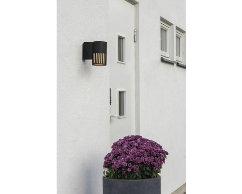 Modern svart utomhusvägglampa på en vit fasad bredvid en dörr, med vertikala ljusöppningar.