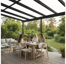 Fyra personer äter vid ett träbord på en träterrass under en svart pergola med tak i en ljus och välskött trädgård.