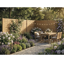 Trädgårdsscen med insynsskydd av trä, trädgårdsmöbler i trä, blommande växter som lavendel och hortensior samt ljusslingor på en uteplats.