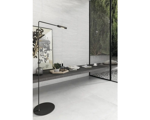 Modernt vardagsrum med strukturerat vitt väggkakel, ljusa golvplattor, en mörkgrå bänk och en minimalistisk svart golvlampa.
