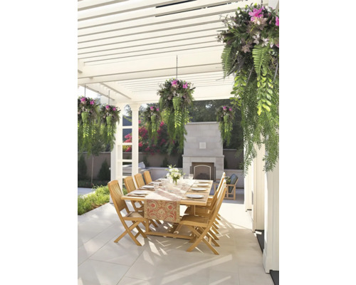 Matplats utomhus under en vit pergola med träbord, trästolar, hängande växter och en eldstad i sten på en stenlagd uteplats.