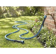 Textil trädgårdsslang ansluten till en utomhuskran på en tegelvägg, liggande på en gräsmatta i en trädgård.