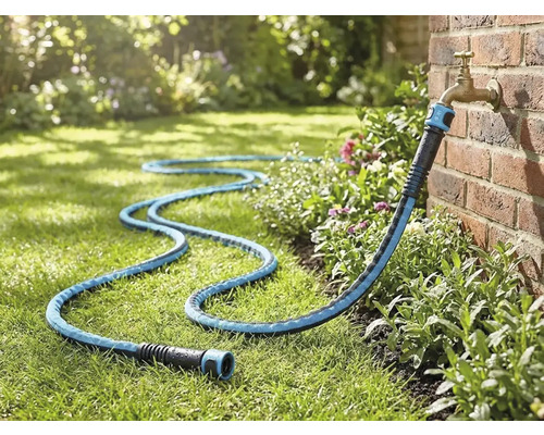 Textil trädgårdsslang ansluten till en utomhuskran på en tegelvägg, liggande på en gräsmatta i en trädgård.