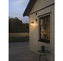 Utomhusvägglampa på en träfasad bredvid ett fönster i skymning, trädäck med ett litet bord.