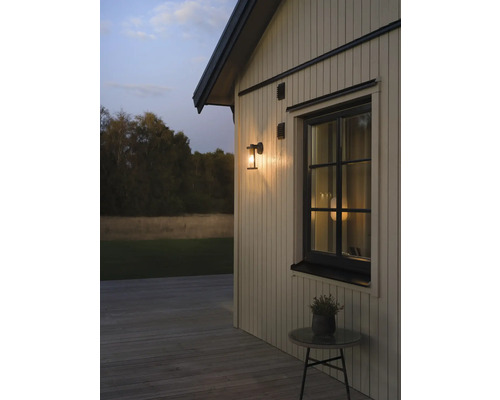 Utomhusvägglampa på en träfasad bredvid ett fönster i skymning, trädäck med ett litet bord.