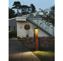 Modern utomhuspollare i cortenstål med rostlook belyser en utemiljö med stenmur och ett modernt bostadshus i skymningen.