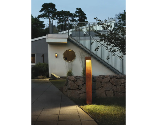 Modern utomhuspollare i cortenstål med rostlook belyser en utemiljö med stenmur och ett modernt bostadshus i skymningen.