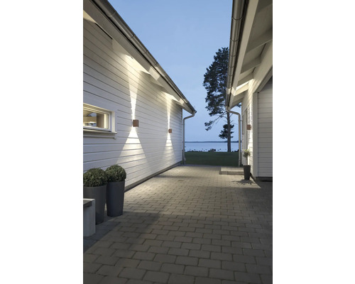 Stenlagd gång mellan vita trähus med moderna fyrkantiga utomhusvägglampor som lyser uppåt och nedåt. Utsikt över en sjö.