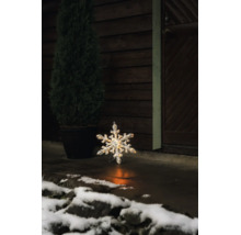 Lysande LED-snöflinga för utomhusbruk på ett stengolv framför en mörk trävägg med en krukväxt och snö.