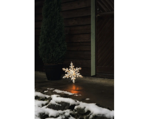 Lysande LED-snöflinga för utomhusbruk på ett stengolv framför en mörk trävägg med en krukväxt och snö.