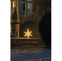 Varmvit LED-snöflinga som utomhusdekoration, placerad på marken i en vintrig trädgård på natten bredvid en buske och ett hus.