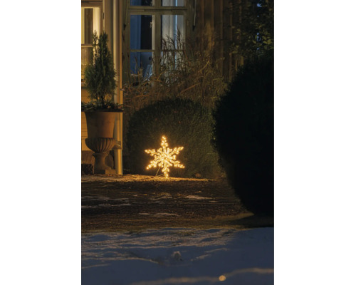 Varmvit LED-snöflinga som utomhusdekoration, placerad på marken i en vintrig trädgård på natten bredvid en buske och ett hus.