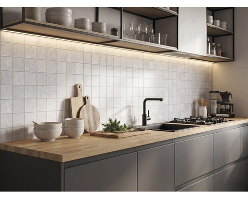 Modern köksmiljö med bänkskiva i trä, mörka skåp, kvadratiskt kakel, svart blandare och belysning under hyllorna.