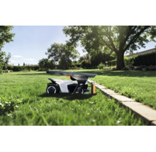 Robotgräsklippare under ett skyddstak på en gräsmatta i en solig trädgård med träd och stenkant.