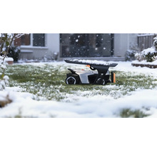 Robotgräsklippare i ett garage på en snöig gräsmatta under snöfall framför ett hus.