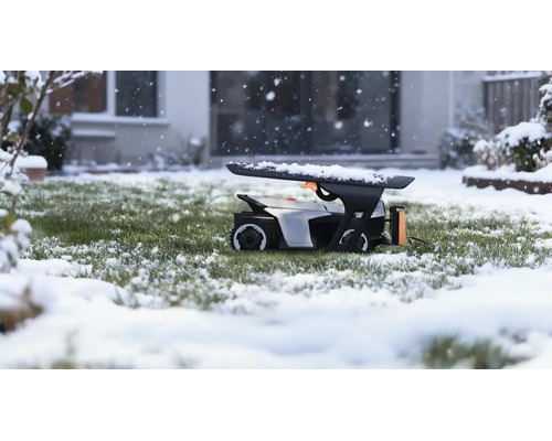 Robotgräsklippare i ett garage på en snöig gräsmatta under snöfall framför ett hus.