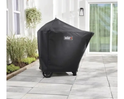 Svart grillöverdrag med Weber Logo på en ljus stenterrass utomhus bredvid växter.